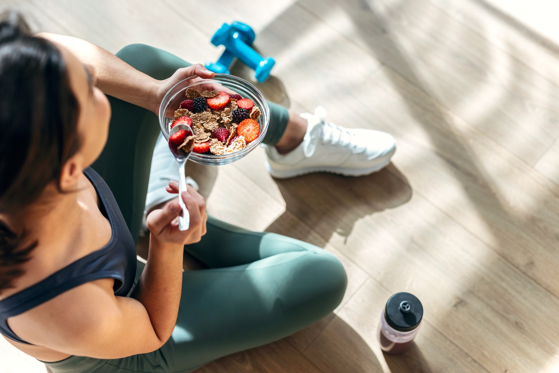 Athletic woman eating a healthy bowl of muesli with fruit sitting on floor in the kitchen at home